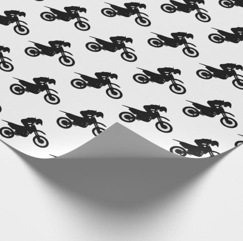 Op de afbeelding: Wit cadeaupapier met een herhaald patroon van zwarte motorfiets silhouetten. Het papier is gedeeltelijk gevouwen en onthult een witte onderkant en een schaduw.