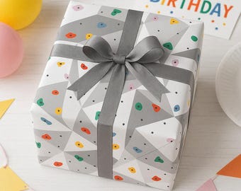 May include: A square gift box wrapped in white paper featuring a gray geometric pattern with small, colorful climbing holds. The box is tied with a dark gray satin ribbon tied in a bow on top. A card with the text HAPPY BIRTHDAY is visible.