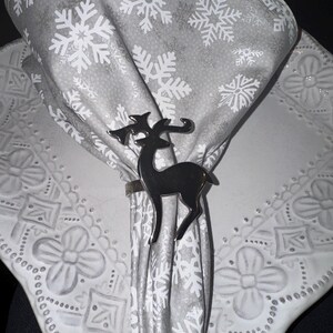 May include: A silver reindeer napkin ring holds a gray napkin with white snowflake patterns. The napkin rests on a white plate with embossed floral designs, creating a festive holiday table setting.