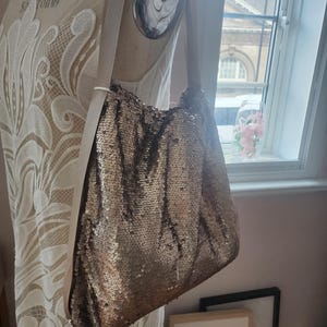 Peut inclure: Un sac à bandoulière de style hobo recouvert de sequins scintillants dans les tons bronze. Le sac présente une large bandoulière lisse de couleur claire fixée aux coins supérieurs du corps souple et non structuré, créant une silhouette froncée.