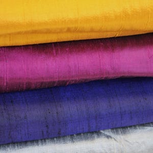 May include: A stack of four folded fabric pieces featuring a distinct slubbed texture. The top layer is bright yellow, followed by a vibrant magenta, a deep royal blue, and a bottom layer of shimmering silver, all displaying a subtle sheen.