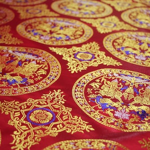 May include: Red fabric with a repeating pattern of gold and blue embroidered circles. Each circle features a stylised design of two mythical creatures with wings and tails. The fabric has a subtle sheen and a luxurious feel.