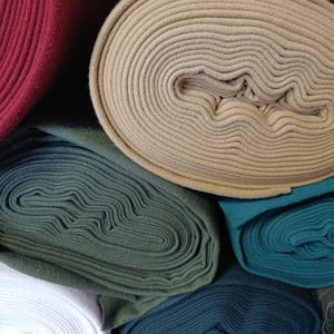 May include: A collection of rolled fabric in various colors, including burgundy, beige, dark green, teal, and navy blue. The fabric appears to be a soft, textured material, possibly fleece or felt, suitable for crafting or sewing projects.