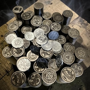 May include: A collection of cylindrical metal wax seal stamps featuring various intricate engravings on their circular faces, including ships, crosses, and heraldic symbols, arranged in a dense cluster with some loose metal coins interspersed.