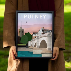 May include: A framed art print featuring the word "PUTNEY" at the top. The artwork depicts a bridge over a river, with buildings and trees in the background. The print is held by a person in a brown coat, with a green grassy background.