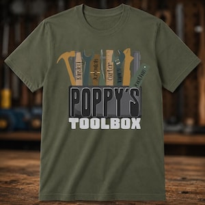 May include: An olive green t-shirt featuring a graphic of a dark gray toolbox with the text "POPPY'S TOOLBOX" in white and black. Inside the toolbox are various tools including a hammer, wrench, and ruler, each personalized with names.