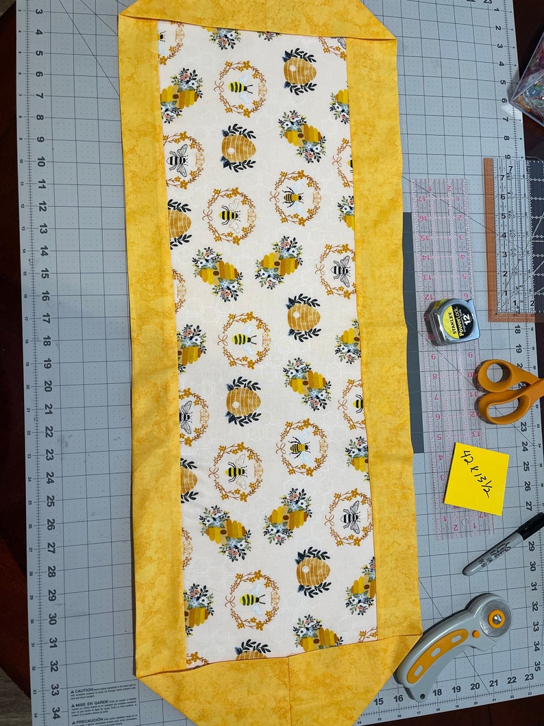 May include: A rectangular table runner with a bee and honeycomb pattern on a cream background, framed by a yellow border. The runner measures approximately 42.5 inches long. The design includes bees, floral accents, and beehives.