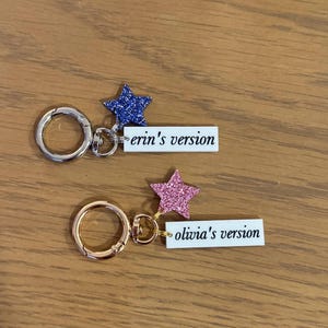 May include: Two keychains with star-shaped glitter accents. One has a blue star and reads "erin's version," the other has a pink star and reads "olivia's version." Both have a metal clasp.