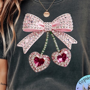 Puede incluir: Camiseta gris oscuro con un lazo rosa y dos cerezas en forma de corazón adornadas con pedrería. El lazo está centrado sobre las cerezas, que cuelgan debajo. La camiseta tiene un aspecto de lavado vintage.