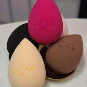 May include: Four makeup sponges in different colors: pink, black, beige, and brown. Each sponge has the word "Daub" written on it.