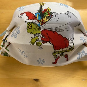 May include: A white face mask with a blue and white snowflake pattern. The Grinch is wearing a Santa hat and carrying a bag of presents. The Grinch is green and has a mischievous expression.