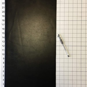 Może przedstawiać: Czarny arkusz materiału o wymiarach 63,5 cm x 48,3 cm, rozłożony na białym tle z siatką. Srebrny długopis leży na siatce po prawej stronie arkusza.