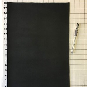 Może przedstawiać: Czarny arkusz pianki do rękodzieła, o wymiarach około 30 cm x 43 cm, leży płasko na białej powierzchni w kratkę. Po prawej stronie arkusza pianki leży przezroczyste pi&oacute;ro.