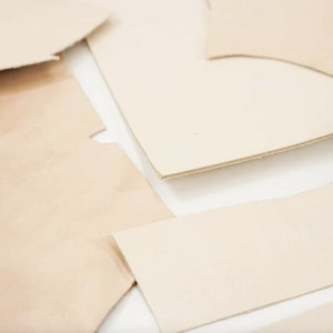 May include: Close-up of several pieces of off-white leather cut into different shapes. The leather is smooth and appears to be high quality.