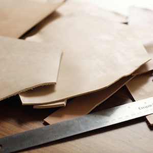 May include: A close-up of several pieces of light brown leather cut into different shapes. A metal ruler with the word "Empire" printed on it is lying on top of the leather.