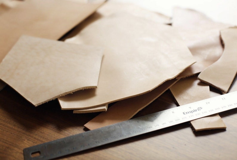 May include: A close-up of several pieces of light brown leather cut into different shapes. A metal ruler with the word "Empire" printed on it is lying on top of the leather.
