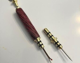 May include: A sewing seam ripper with a red and brown wooden handle and gold-colored metal accents. The seam ripper is next to a matching gold-colored metal cap. A silver chain is attached to the top of the handle.