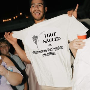 May include: A white t-shirt with black text that reads "I got Sauced at Cameron & Maggie's Wedding". The text is accompanied by a graphic of a fork and spaghetti.