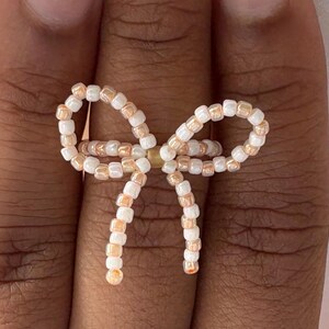 May include: A beaded ring in the shape of a bow. The ring is made of small, round beads in shades of white and peach. The bow design is centered on the ring, creating a delicate and feminine accessory.