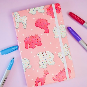 Puede incluir: Una libreta rosa con un cierre de banda elástica blanca. La portada presenta un patrón repetido de galletas con forma de animales con chispas de colores. Las galletas tienen forma de caballo, elefante, jirafa y rinoceronte.