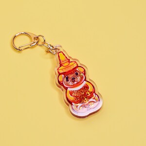 May include: A clear keychain with a gold metal ring. The keychain features a cartoon bear shaped like a honey bottle with a red and yellow lid. The bear is filled with gold glitter.