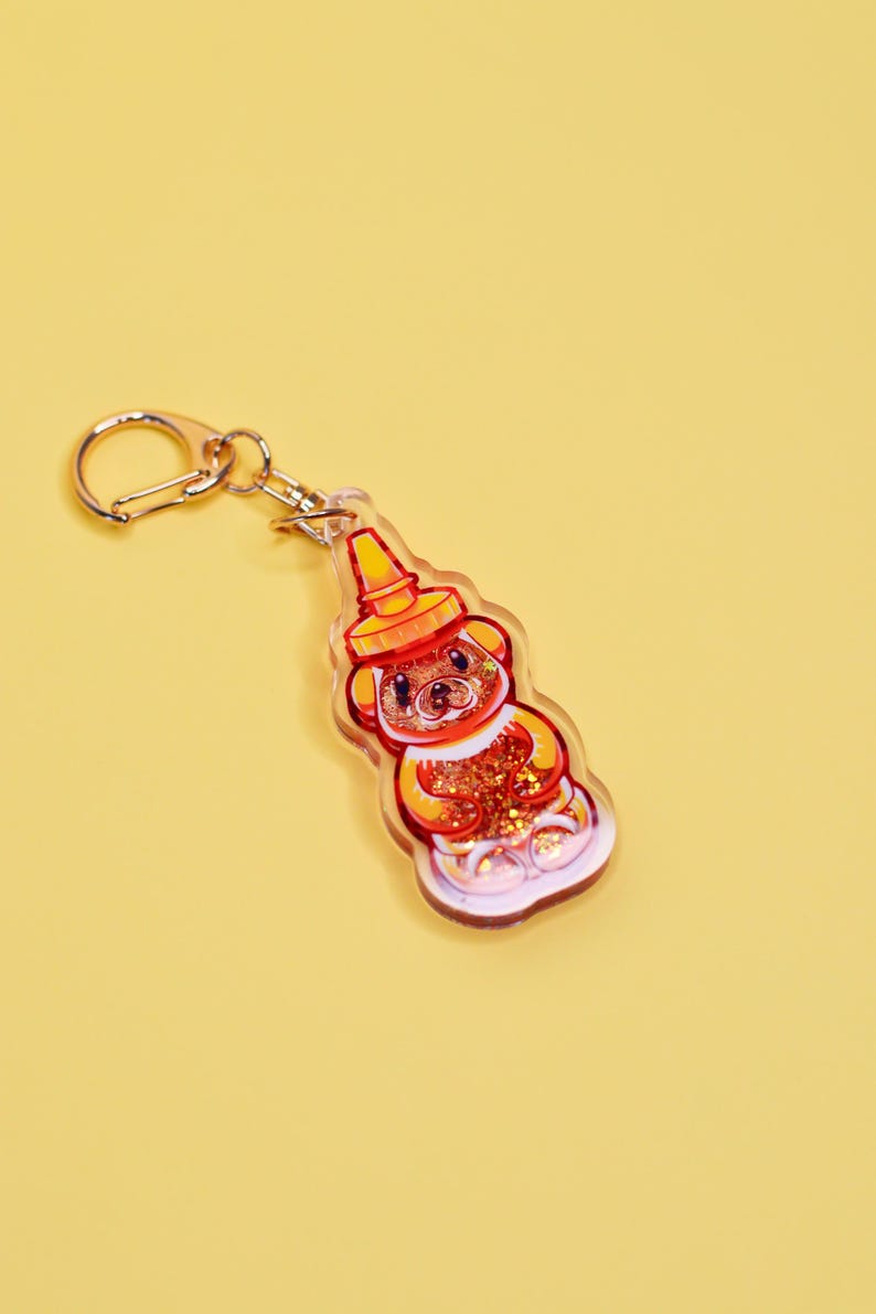 May include: A clear keychain with a gold metal ring. The keychain features a cartoon bear shaped like a honey bottle with a red and yellow lid. The bear is filled with gold glitter.