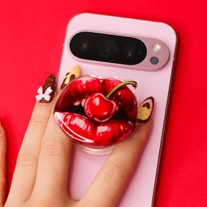 Puede incluir: Un smartphone rosa con un agarre emergente con un diseño de cereza roja y labios. El teléfono se sostiene contra un fondo rojo brillante. El agarre emergente es redondo y transparente.