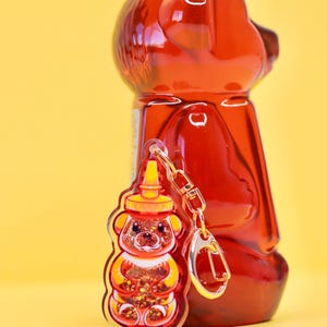 May include: A red and yellow bear-shaped honey bottle keychain with glitter inside. The keychain is hanging from a gold chain and is next to a larger red honey bottle.