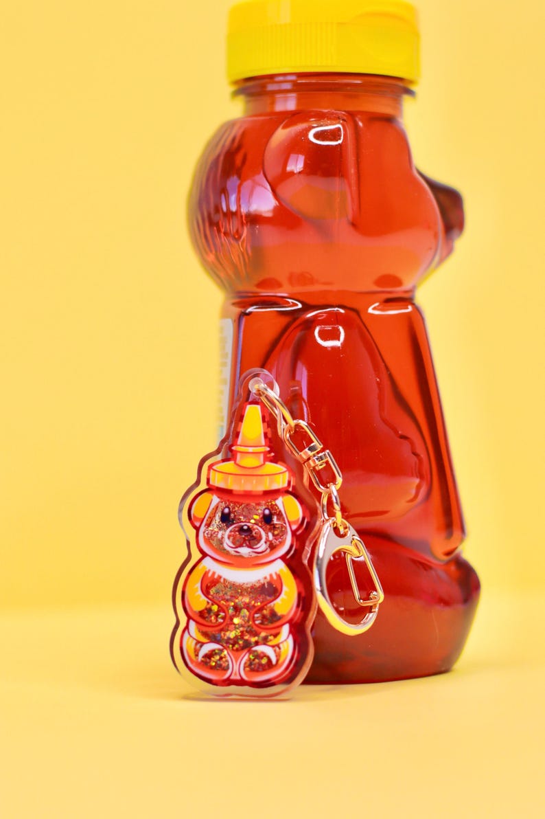 May include: A red and yellow bear-shaped honey bottle keychain with glitter inside. The keychain is hanging from a gold chain and is next to a larger red honey bottle.