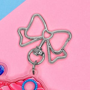 以下が含まれることがあります： 銀色のリボン型のキーホルダーチャーム。チャームは金属製で、中央にハートが付いたリボンのデザインが特徴です。リングと、ピンクと青のデザインが施された透明なプラスチック片に取り付けられています。