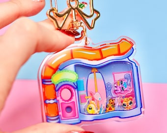 Op de afbeelding: Een transparante plastic sleutelhanger in de vorm van een klein huis met een gouden strik bovenop. Het huis heeft een roze deur en ramen, en er zitten kleine cartoondieren in.