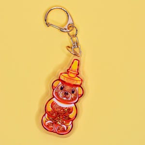 May include: A gold keychain with a clear acrylic charm shaped like a honey bear bottle. The bear is filled with glitter and has a red and orange color scheme.