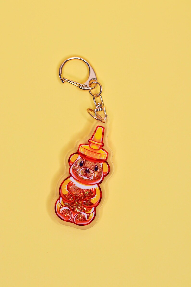 May include: A gold keychain with a clear acrylic charm shaped like a honey bear bottle. The bear is filled with glitter and has a red and orange color scheme.