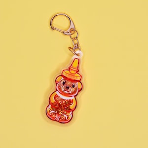 May include: A gold keychain with a clear plastic charm shaped like a honey bear. The bear is filled with glitter and has a red and yellow design.