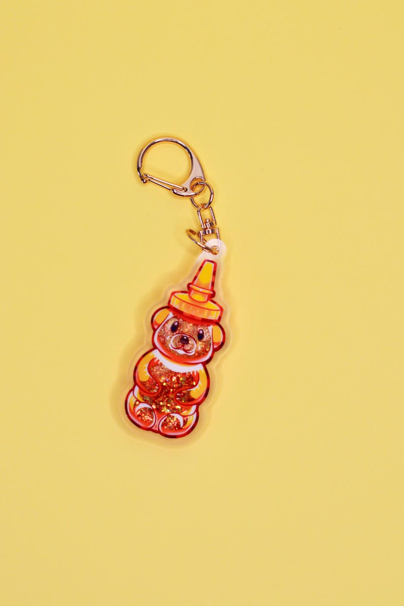 May include: A gold keychain with a clear plastic charm shaped like a honey bear. The bear is filled with glitter and has a red and yellow design.