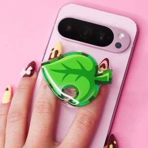 May include: A green leaf-shaped phone grip is attached to a pink smartphone. The grip has a glossy finish and a detailed leaf design. The background is a solid pink color.