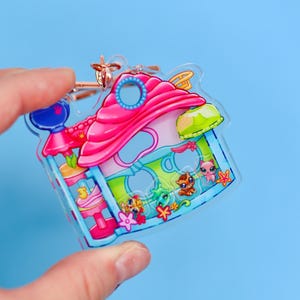 以下が含まれることがあります： ピンクの屋根と青い縁取りの家型をした、透明なアクリルキーホルダー。カラフルな漫画のキャラクターと花のアクセントが特徴です。ゴールドカラーのキーリングが付いています。