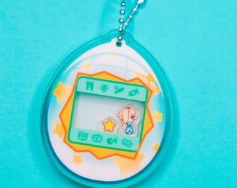 Puede incluir: Un llavero de plástico azul y amarillo con forma de mascota digital con un personaje de dibujos animados en la pantalla. El llavero tiene una cadena plateada.