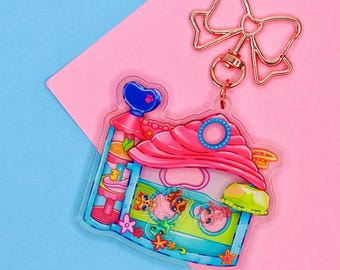 May include: A clear acrylic keychain featuring a pink and blue house design with cartoon animal characters inside. The keychain has a gold-colored bow-shaped clasp and a ring for attaching keys. The background is a pink and blue color block.