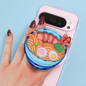Puede incluir: Un soporte para teléfono de acrílico transparente con un diseño de cuenco de ramen colorido. El cuenco tiene rayas azules y blancas y está lleno de fideos, carne, verduras y un huevo duro. El soporte está adherido a un teléfono rosa.