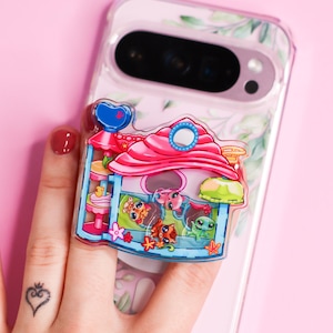 May include: A clear phone case featuring a botanical leaf pattern and a colorful acrylic phone grip shaped like a multi-level pet playhouse. The grip displays cartoon animals, including cats and a turtle, within a blue structure with a pink roof.