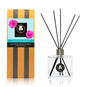 May include: A glass bottle reed diffuser with black reeds and a gold cap. The bottle has a label with a pink hibiscus flower and the text "RAINIER'S GIFTS CELEBRATE LIFE SOUTH PACIFIC HIBISCUS REED DIFFUSER | DIFFUSEUR DE PARFUM 6.5 fl oz e 192 ml."