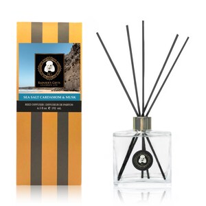 May include: A clear glass bottle with black reeds and a gold lid. The bottle is filled with a clear liquid and has a label that reads "Sea Salt Cardamom & Musk" and "Rainier's Gifts Celebrate Life". The bottle is sitting on a white surface.