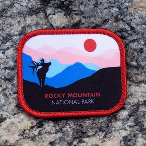 May include: A red, white, and blue embroidered patch with a silhouette of an elk standing in front of a mountain range with a red sun setting behind it. The text "Rocky Mountain National Park" is printed in black at the bottom of the patch.