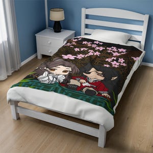 Puede incluir: Una cama individual blanca con una manta de forro polar con estampado en blanco y negro que presenta una pareja de dibujos animados sentada bajo un cerezo en flor. La manta tiene un borde verde y blanco.