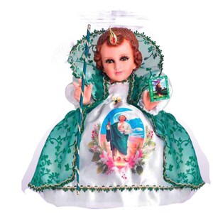 May include: A small, white satin doll dressed in a green lace gown with a white lace trim. The doll has a crown on its head and is holding a staff. The doll is wearing a green and white printed dress with a picture of a saint on it.