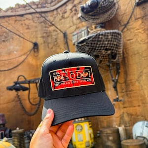 May include: Black baseball cap featuring the Star Wars Episode I: The Phantom Menace logo in red and white. The cap has a curved brim and a structured crown. The background includes a themed environment with droids and other Star Wars elements.