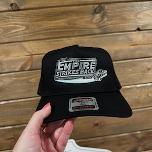May include: Black baseball cap featuring the text "THE EMPIRE STRIKES BACK" with a Star Wars logo. The cap has a flat brim and a mid-profile design. The cap is made of a durable fabric.