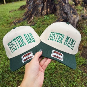 May include: Two baseball caps with the text "FOSTER DAD" and "FOSTER MAMA" embroidered in green on the front. The caps have a cream-colored crown and a dark green brim. The caps are held up in front of a green grassy background.
