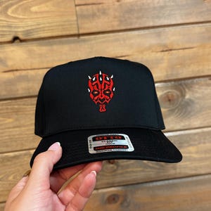 May include: Black baseball cap with a red and white graphic on the front. The graphic appears to be a stylized face. The cap has a curved brim and a structured fit. The cap is being held by a person.